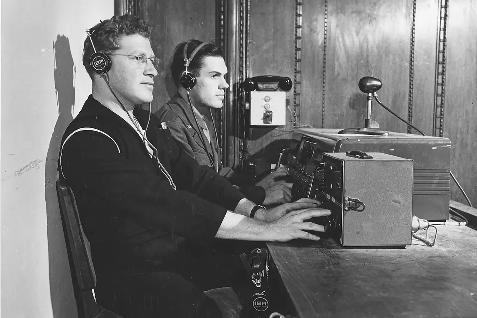 Two men wearing headphones sit at a desk with microphones and an amplifier, working as interpreters in a black-and-white photograph